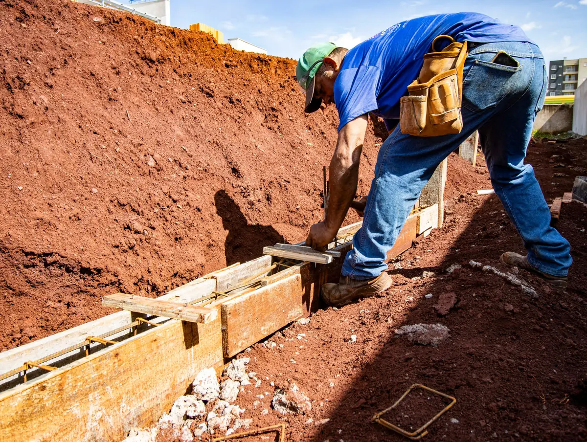 Worker beside wooden footing forms with steel rebar in a clay trench for poured concrete