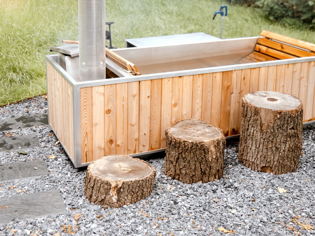 Wood-clad outdoor hot tub with a stove pipe on a bed of grey crushed gravel, with three tree stumps and stone pavers beside it