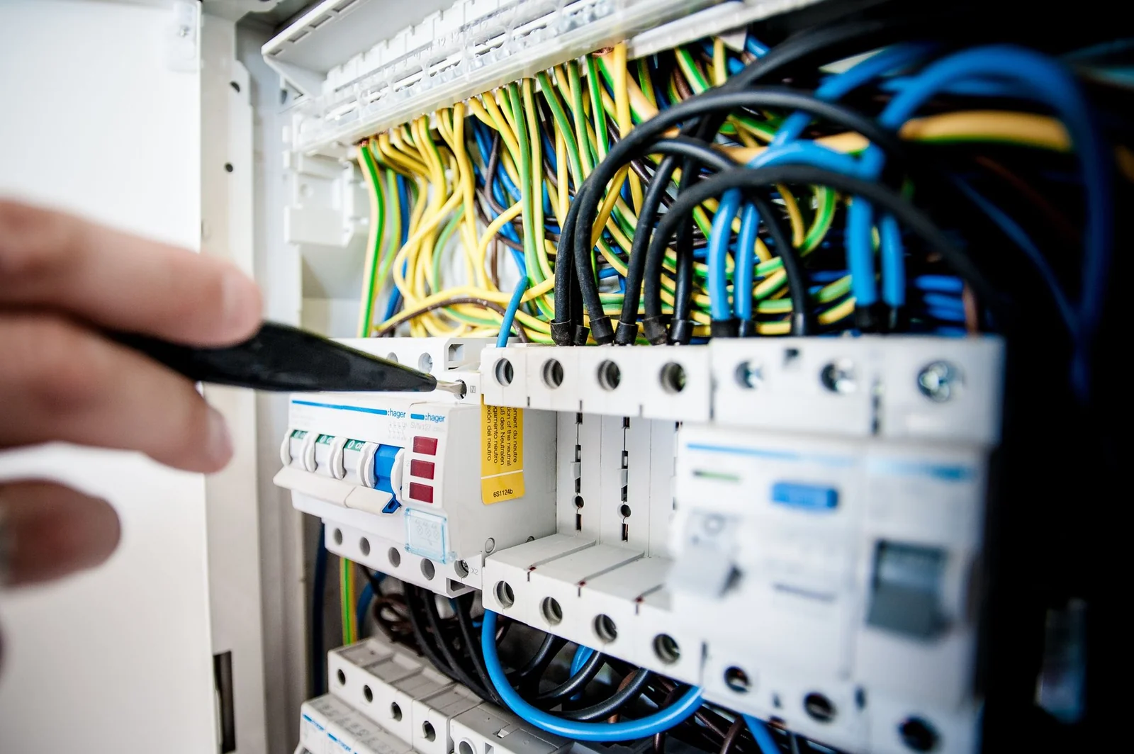 Hand holding a tool at screw terminals on an open circuit breaker panel with color-coded wires