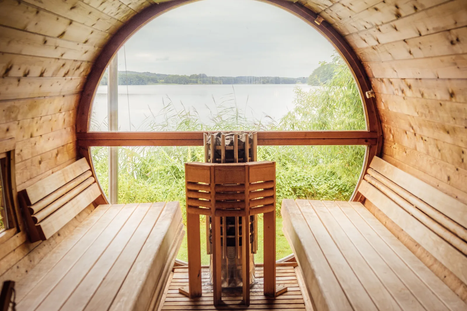 Barrel sauna interior with side benches, a stone-filled pillar heater and a large round window onto a lake