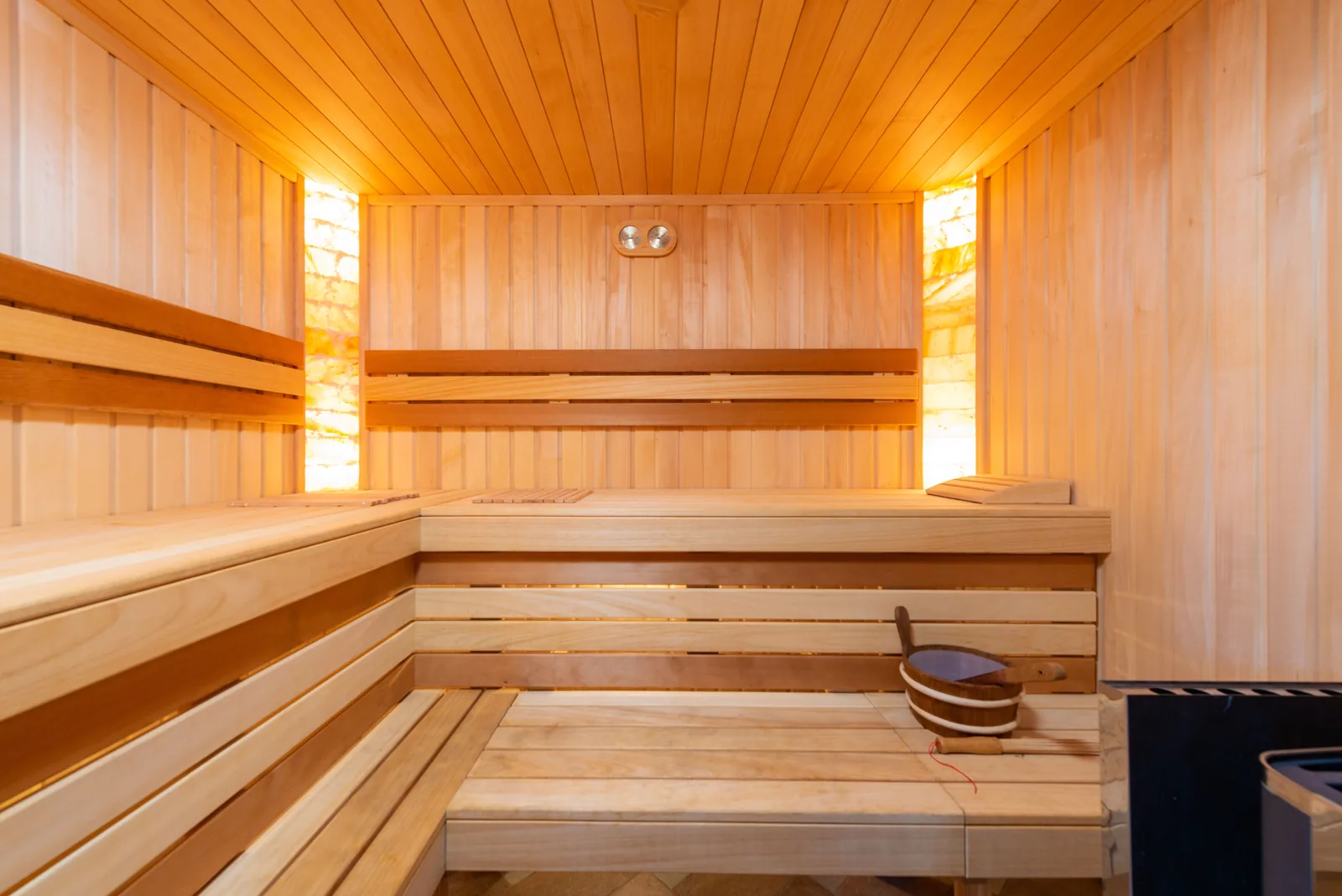 Empty wooden sauna interior with two tiers of benches, a wooden bucket and ladle, and a thermometer and hygrometer on the back wall