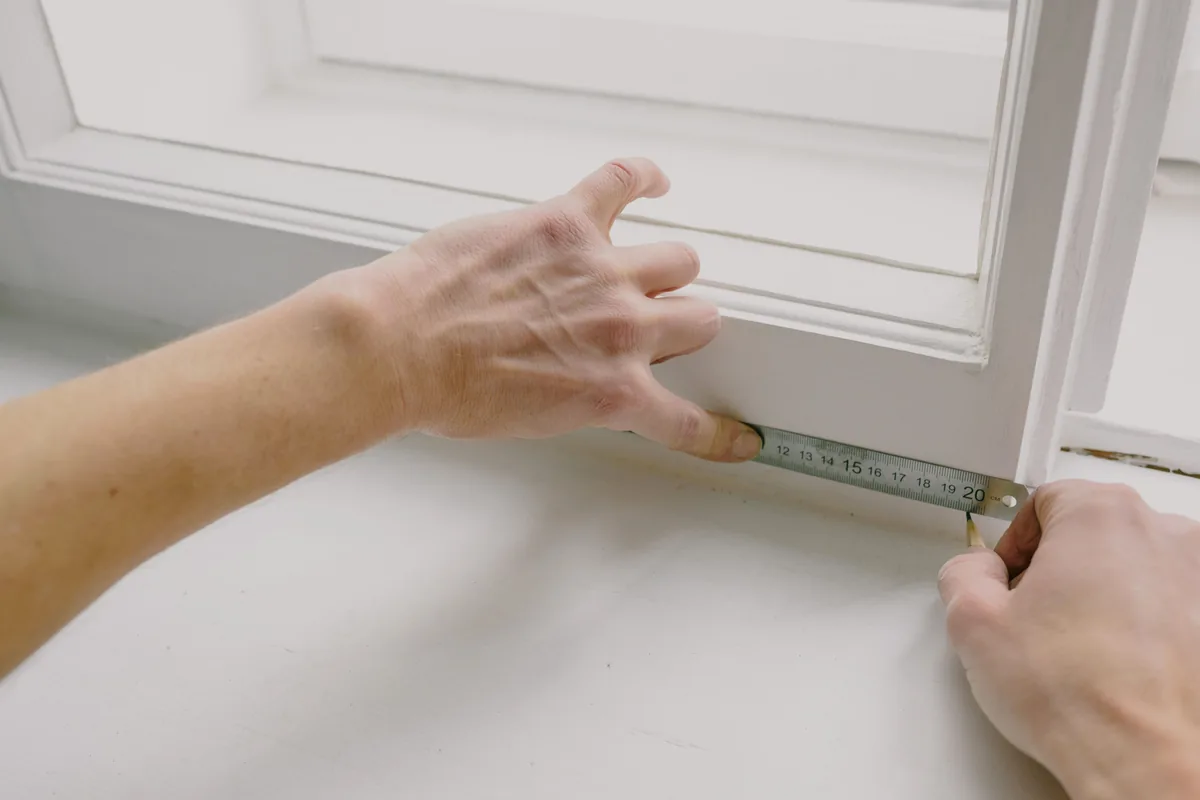 Hands measuring a white window frame sill with a metric ruler and a pencil