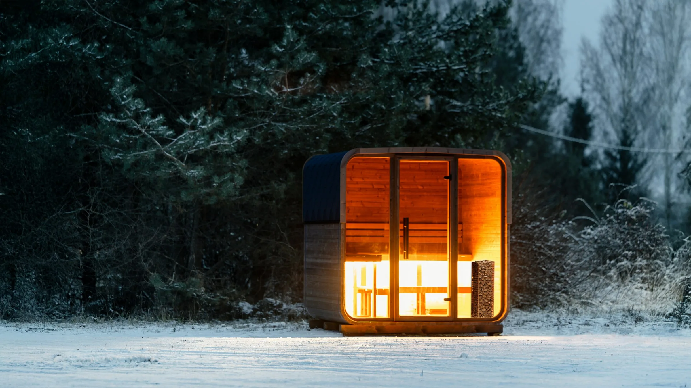 Modern wood-clad sauna with a glass front glowing orange in a snowy forest at dusk.