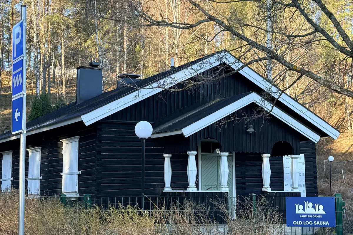 Dark log sauna cabin with a white porch trim and two roof chimneys, behind a green fence in spring