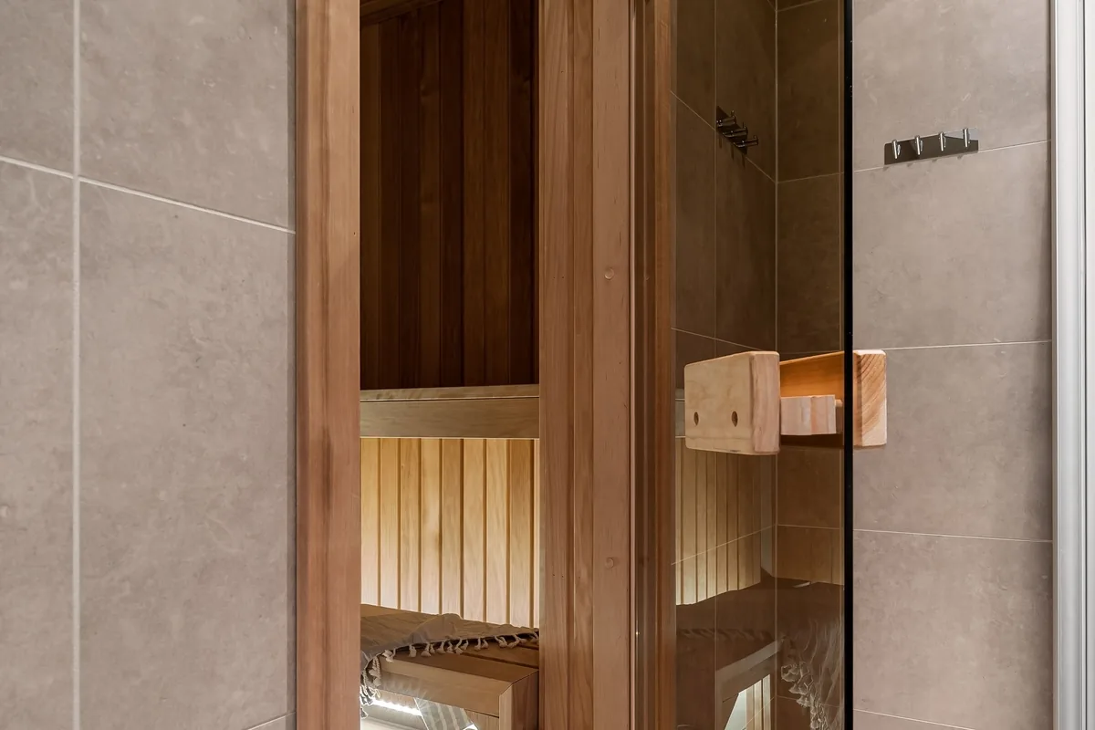 Open glass sauna door in a tiled bathroom, with a view of the wooden bench inside
