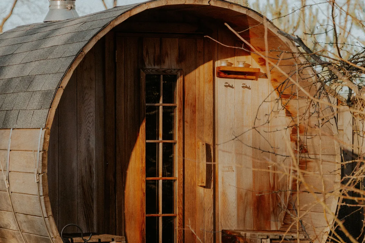Wooden barrel sauna with a shingled roof and a multi-pane glass door, seen through bare branches