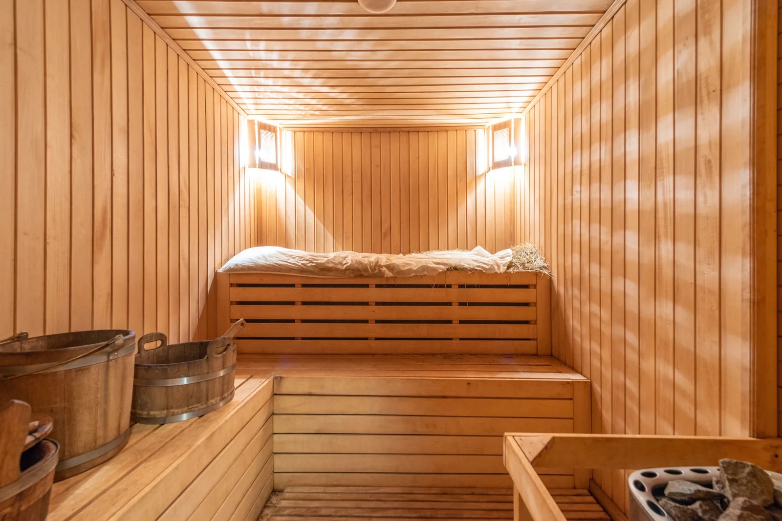 Wooden sauna interior with two tiers of benches, wooden buckets and a stone-filled heater